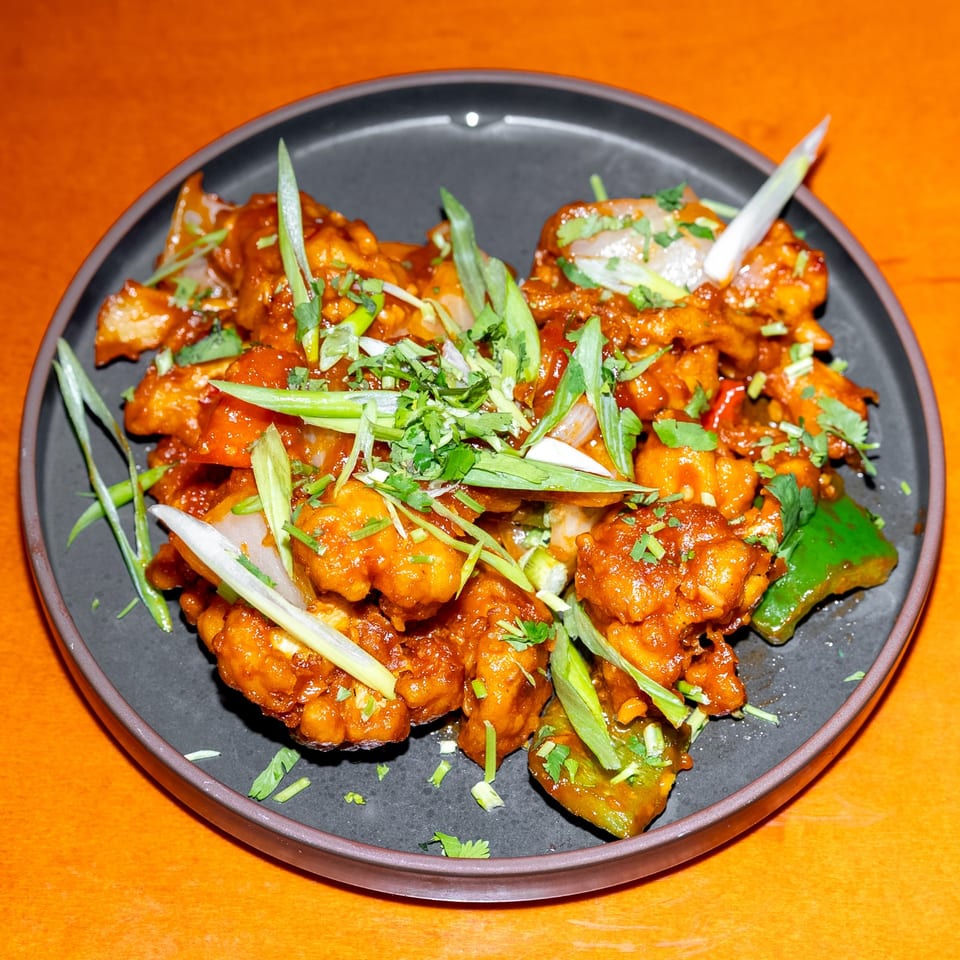 Glazed orange chicken pieces topped with scallions, cilantro, onions, and peppers on a dark plate.